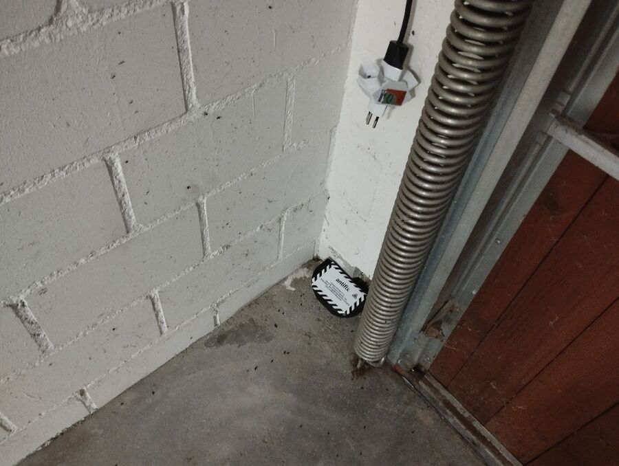 In einer Garage in Cham (6330, Kanton Zug), ist eine schwarze Köderbox am Boden neben der Wand und einem Metallrohr platziert.